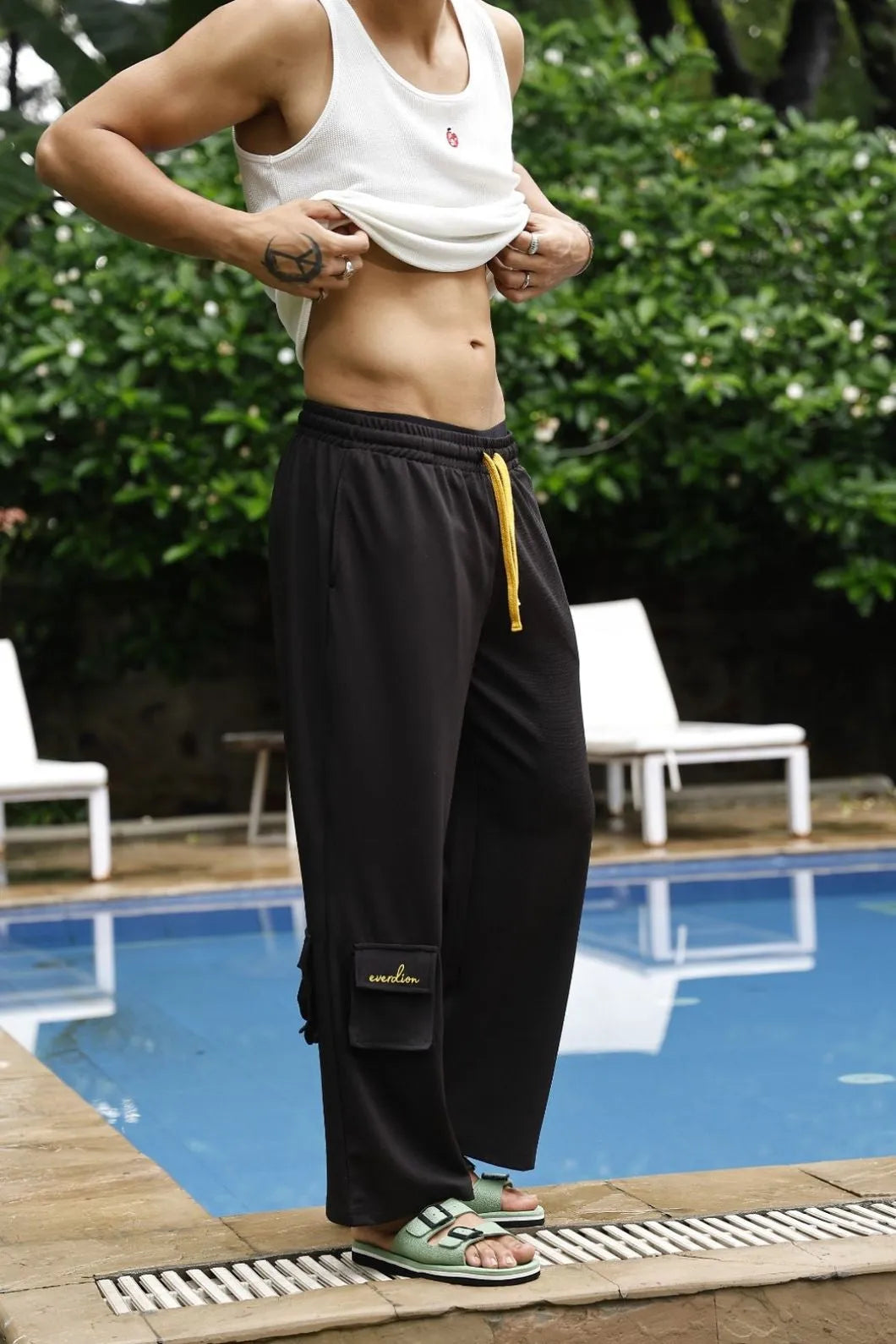 Person wearing a white tank top and black cargo pants by a poolside.