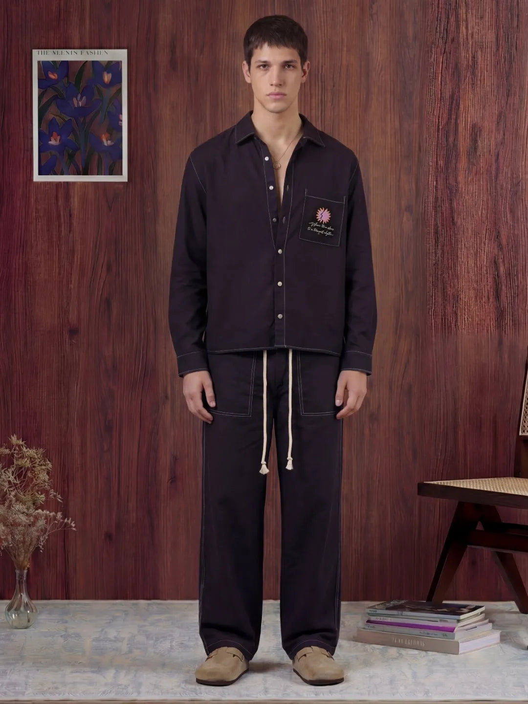 Man wearing a black shirt and pants standing in a room with wooden walls and a small table.