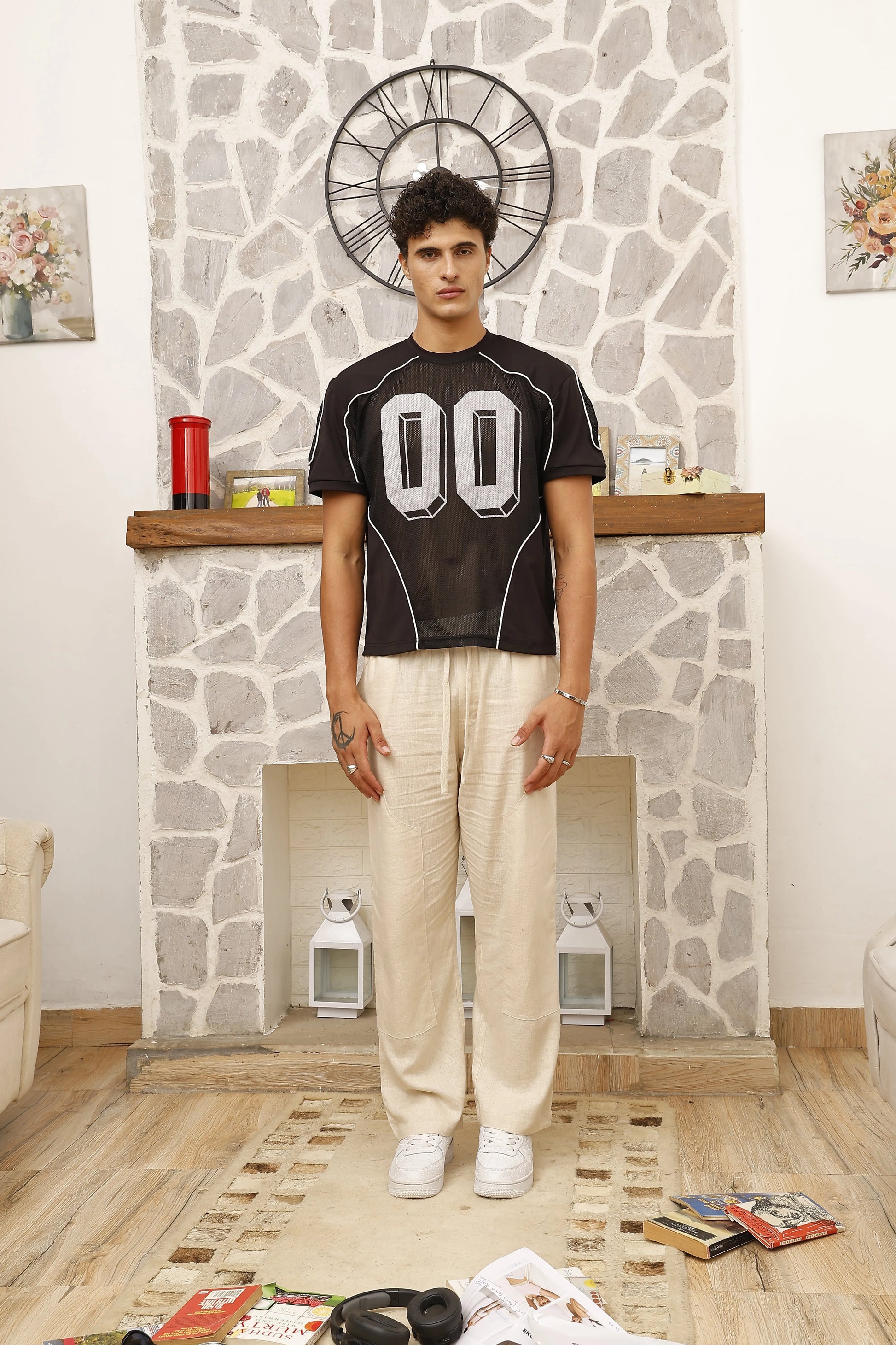 Man wearing a black t-shirt with large white numbers and beige pants standing in a room with stone wall and wooden floor.