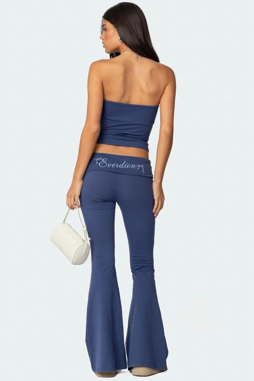 Woman wearing a blue strapless top and flared pants with 'Everdion' branding, holding a white handbag on a light grey background.