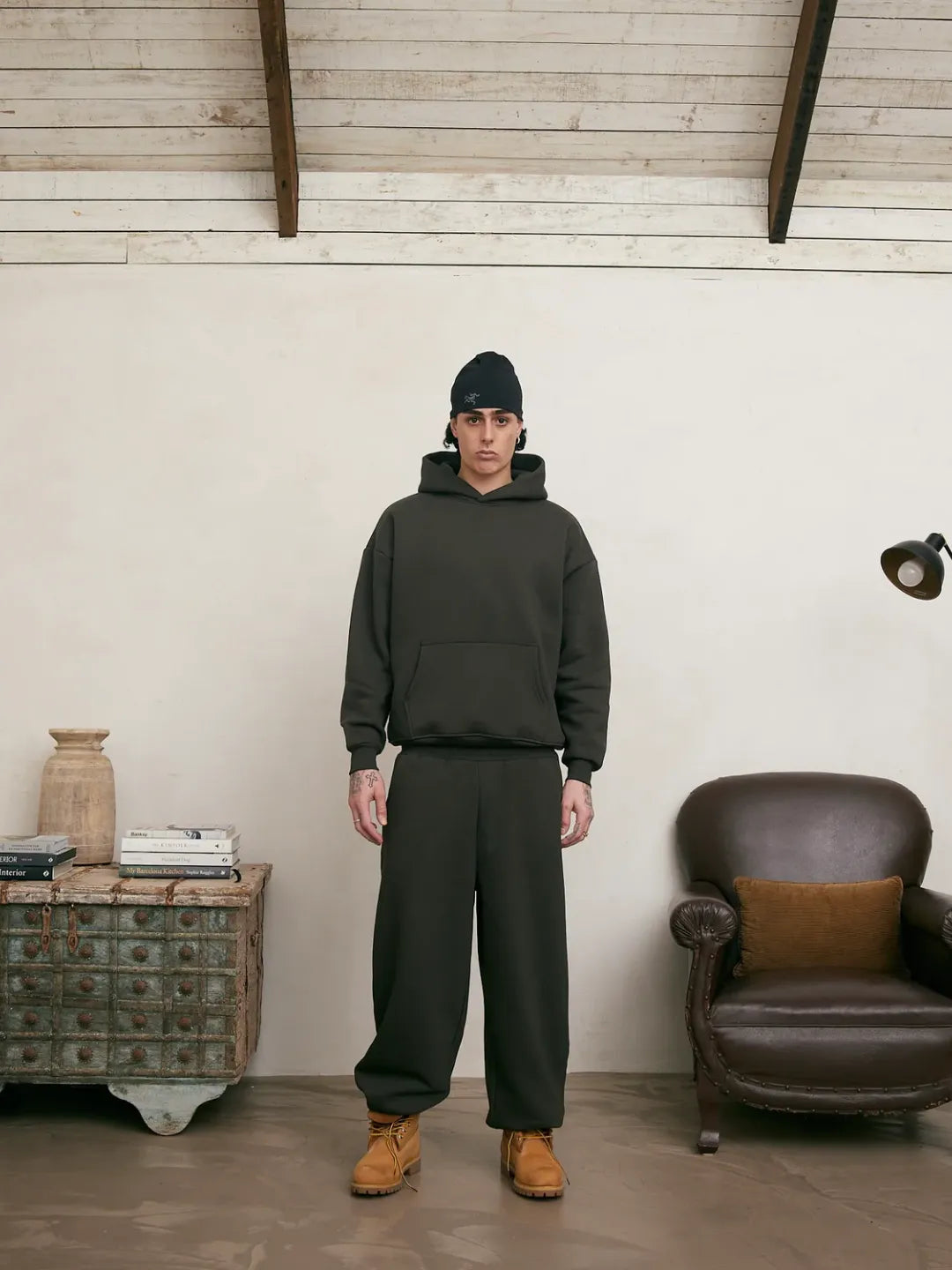 Person wearing a green hoodie and pants standing in a room with a wooden cabinet and leather chair.