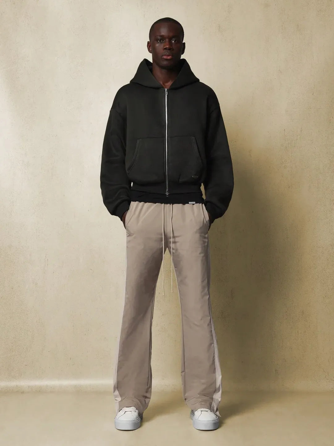 Man wearing a black hoodie and beige pants against a beige wall