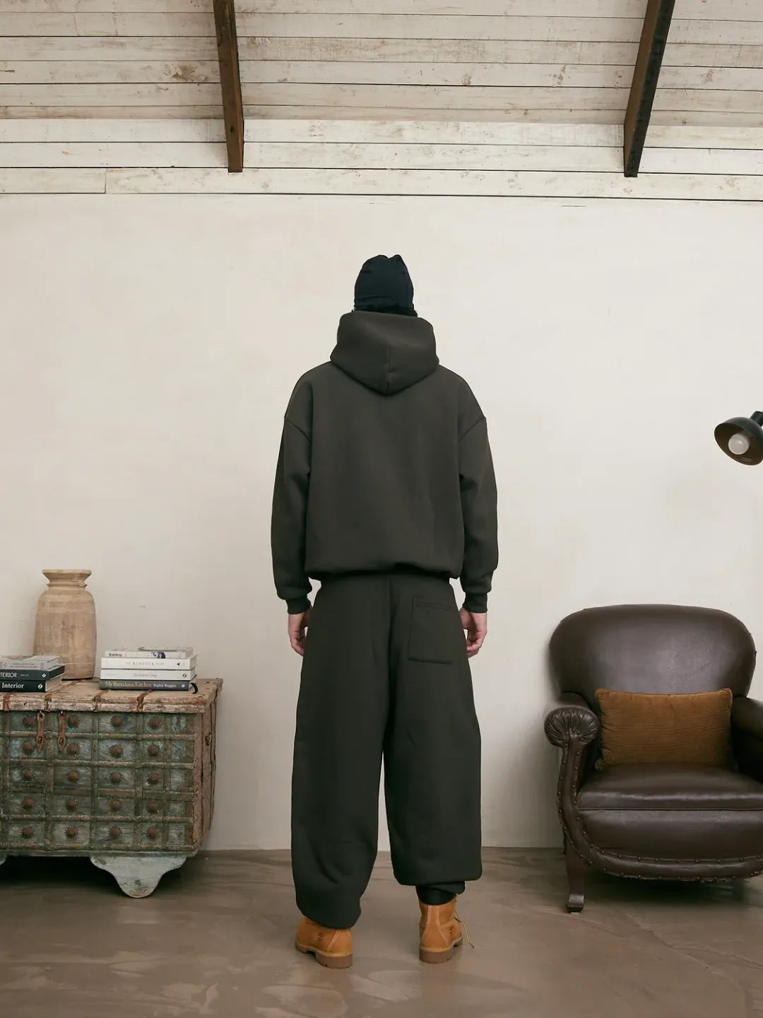 Person wearing a dark green hoodie and pants standing in a room with a wooden cabinet and leather chair.