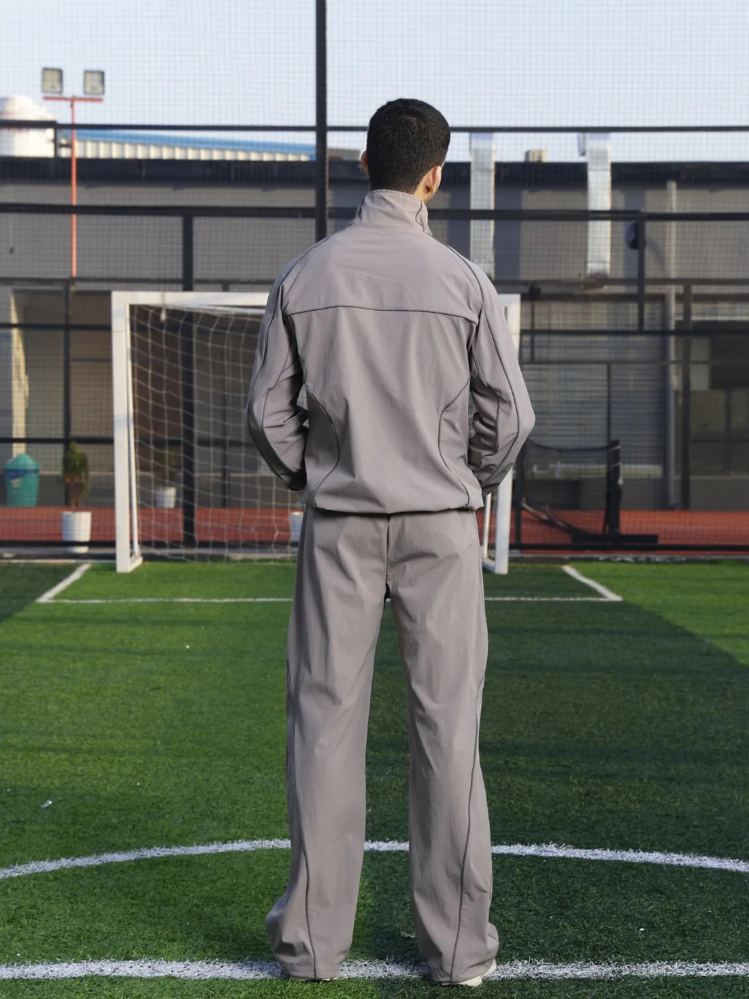 Person wearing a grey tracksuit standing on a soccer field with a goalpost in the background.