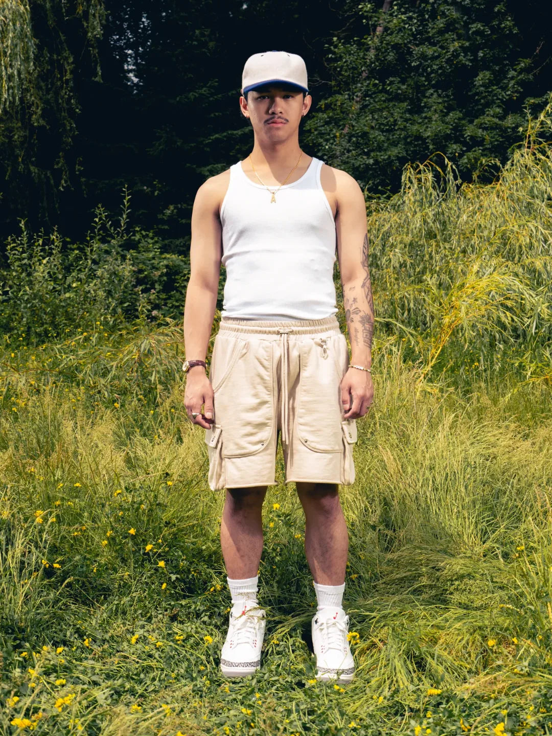 Man standing in a grassy field wearing a white tank top, beige shorts, and a cap.