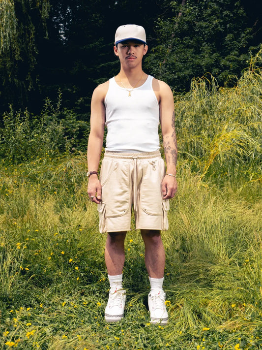 Man standing in a grassy field wearing a white tank top, beige shorts, and a cap.