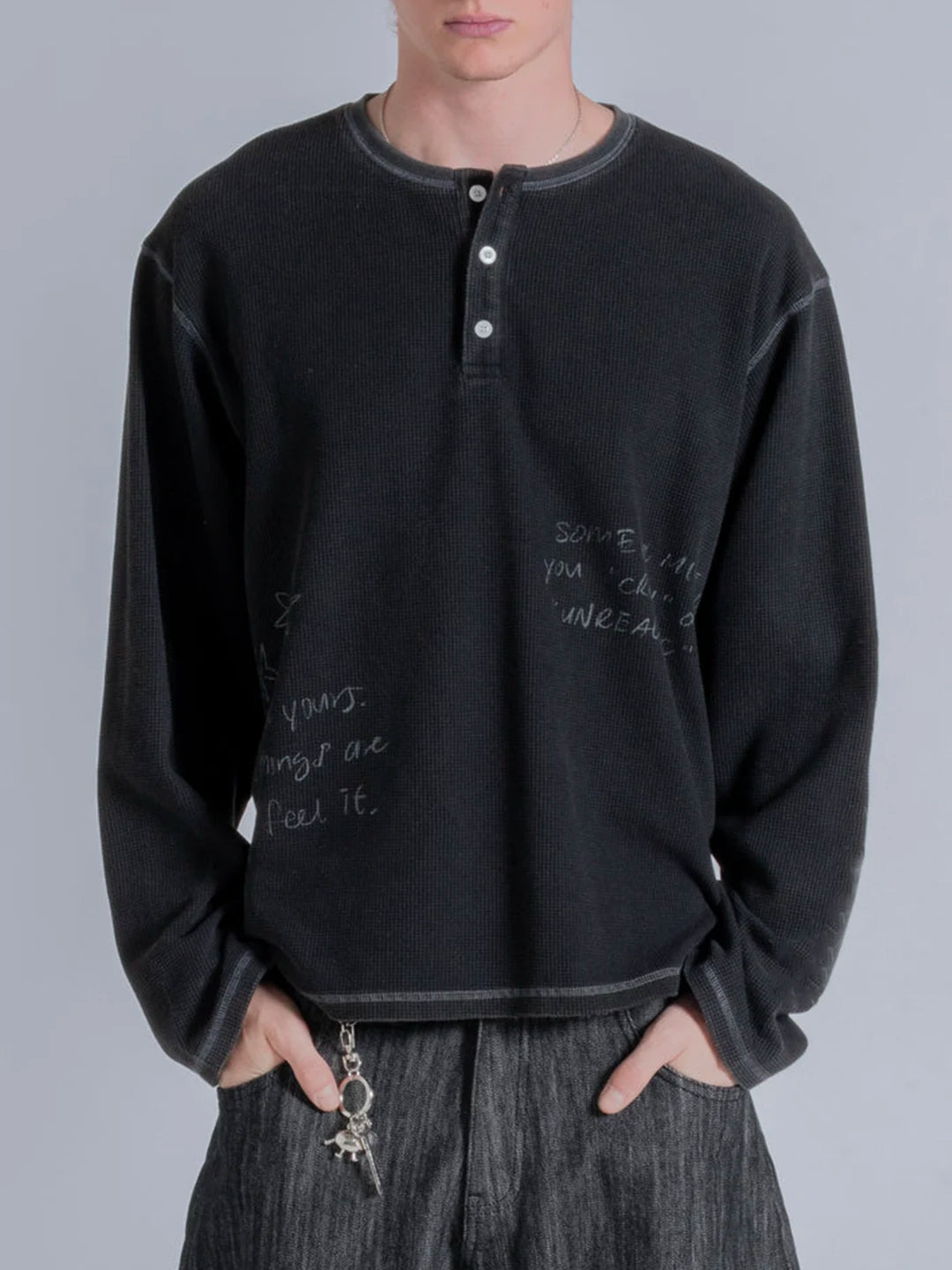 A person wearing a faded black long-sleeve henley t-shirt with white handwritten-style text graphics on the front, paired with dark gray pants