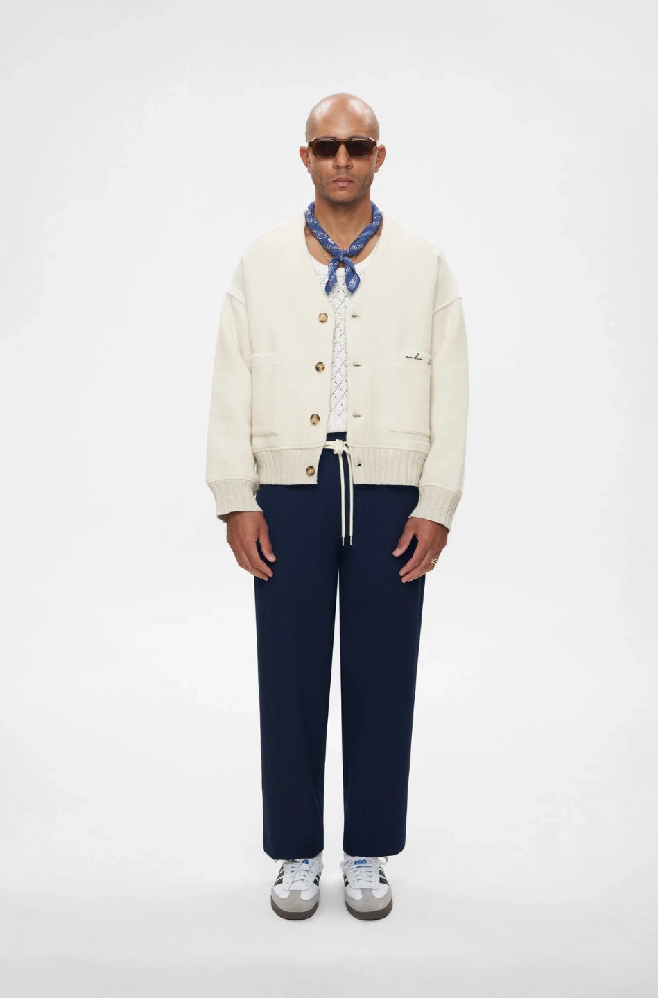 Man wearing a cream cardigan, navy pants, and sunglasses on a white background