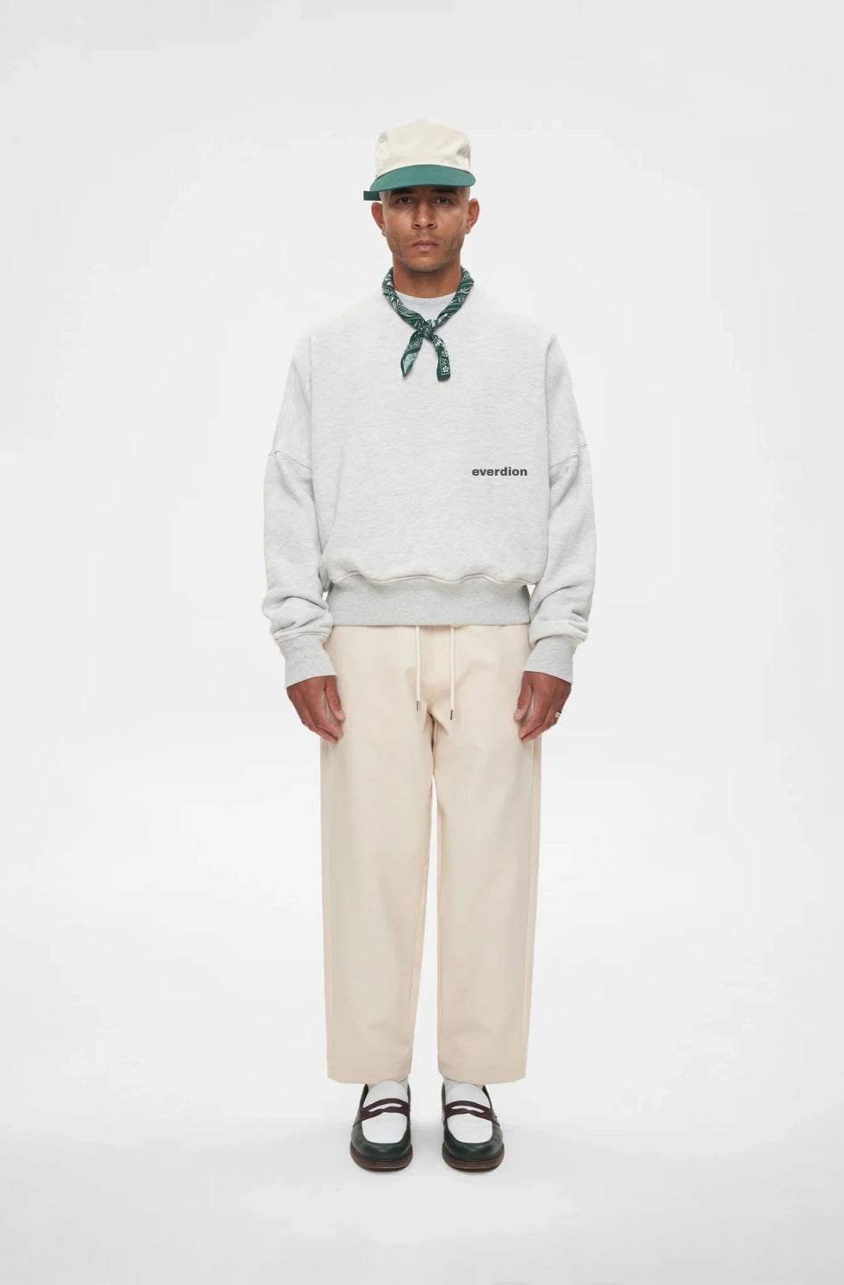 Person wearing a light grey sweatshirt with text, beige pants, and a cap on a white background
