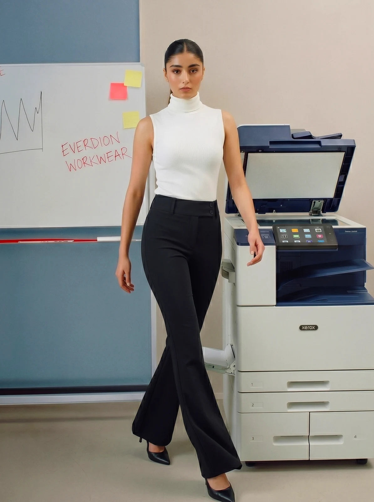 Woman in a white top and black pants standing next to a printer in an office setting.