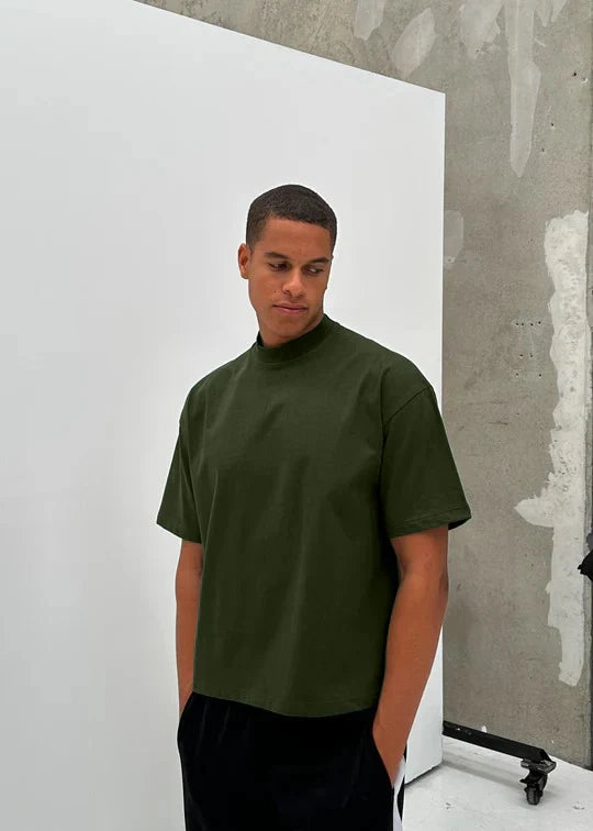 Man wearing a olive t-shirt standing against a white and gray wall.