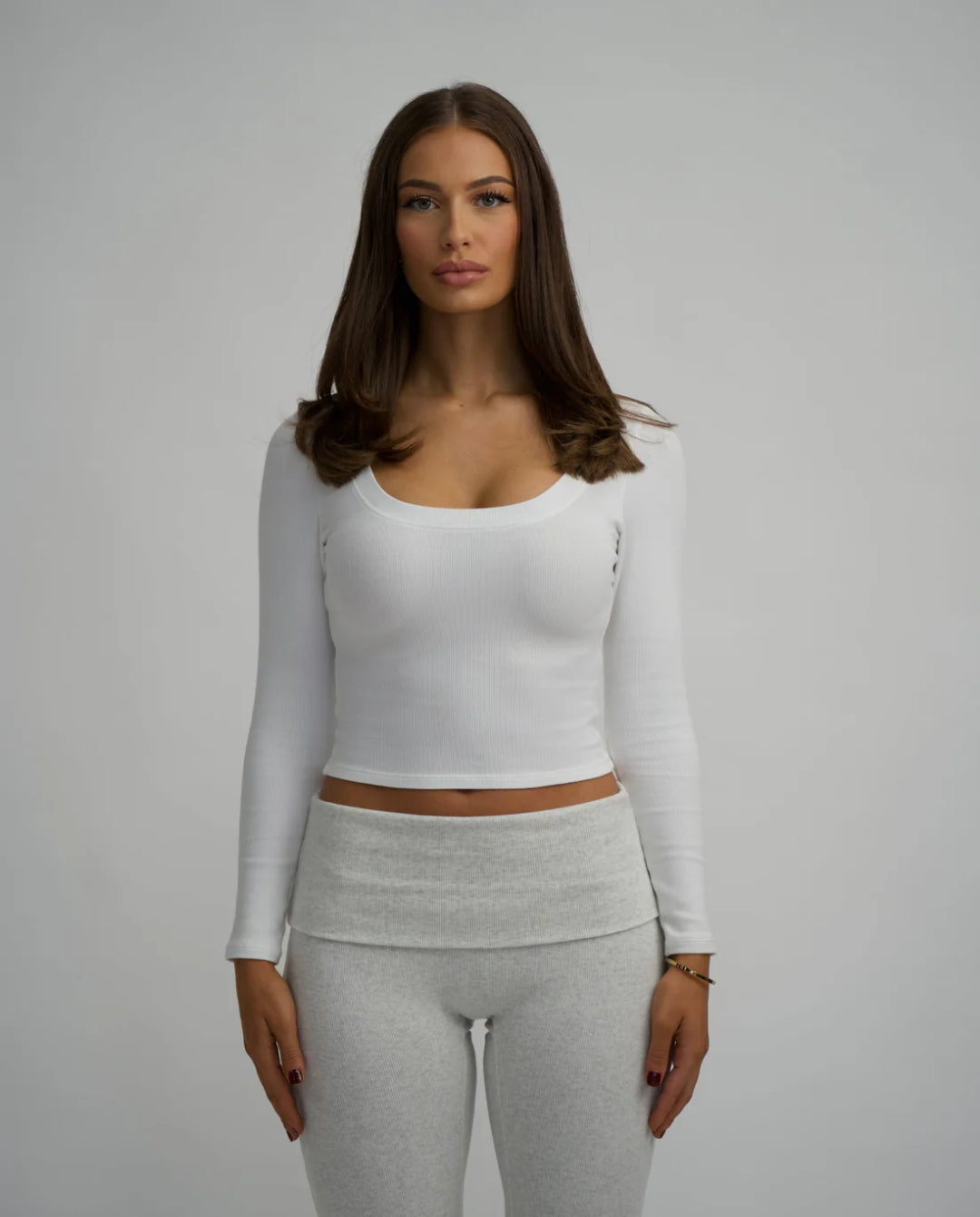 Woman wearing a white long-sleeve top and grey pants on a plain background