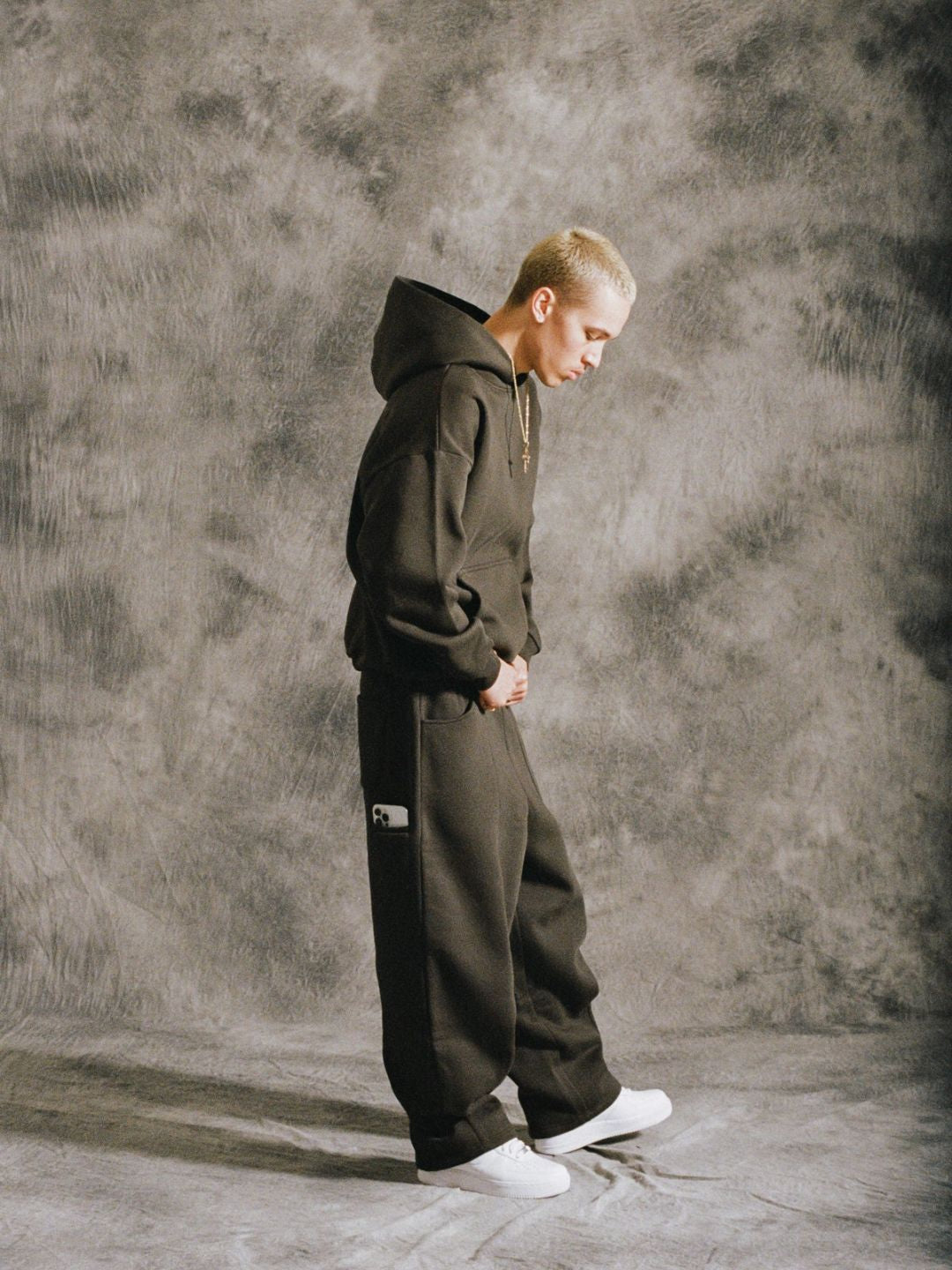 Person wearing a brown hoodie and pants against a textured grey wall.