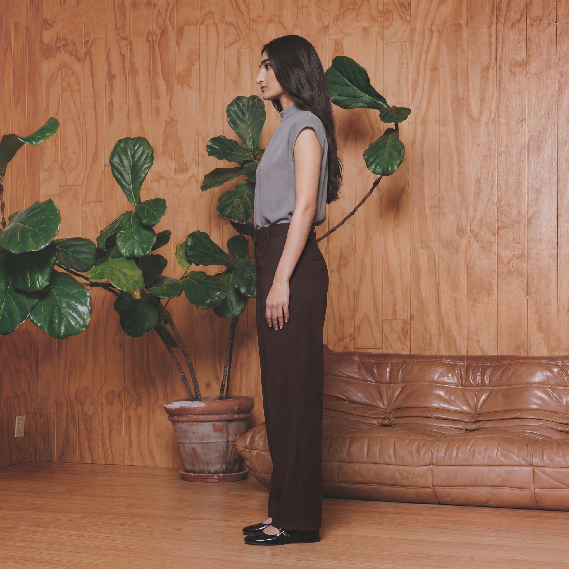 Woman standing next to a potted plant in a room with wooden walls and a brown leather couch.