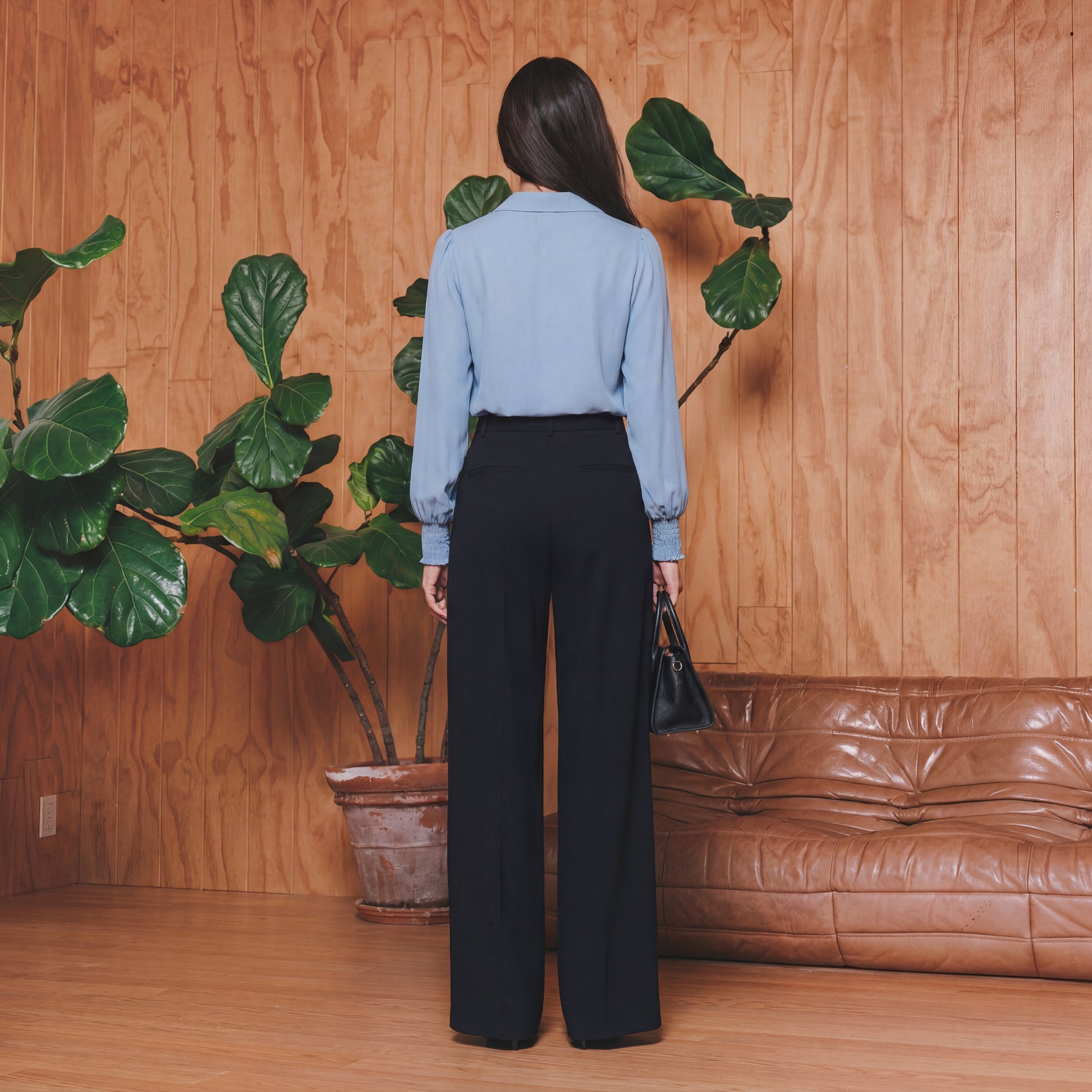 Woman standing in a room with wooden walls, a brown leather couch, and a potted plant.
