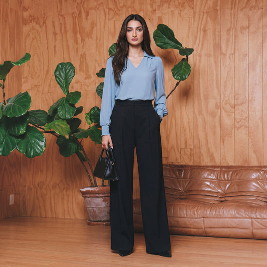 Woman in a blue blouse and black pants standing in a room with wooden walls and a brown leather couch.