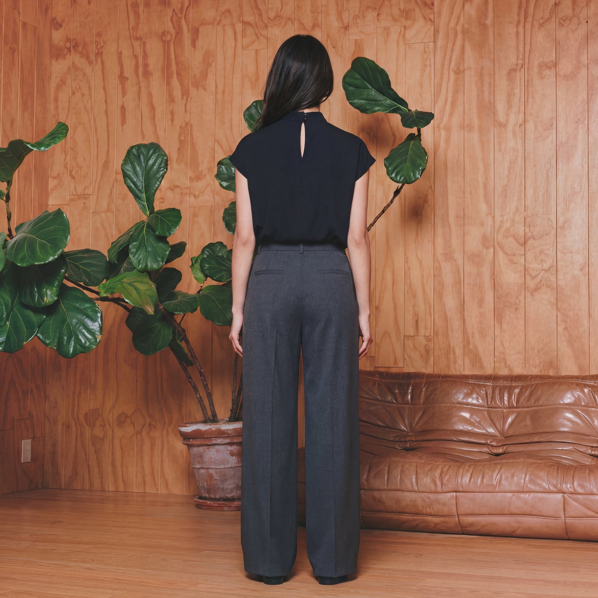 Person wearing a black top and dark pants standing in front of a wooden wall with plants and a brown leather couch.