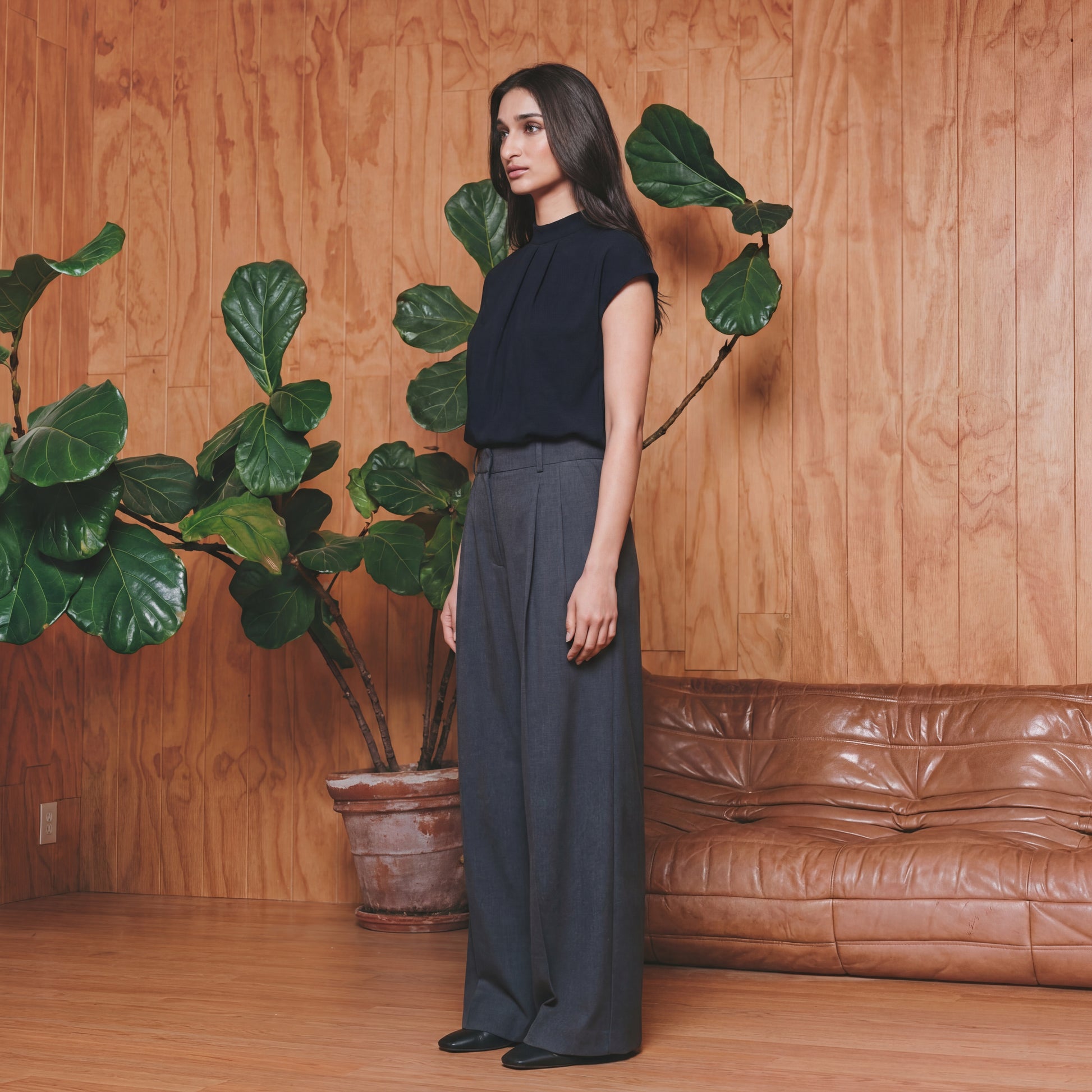 Woman standing in a room with wooden walls, a brown leather couch, and large green plants.