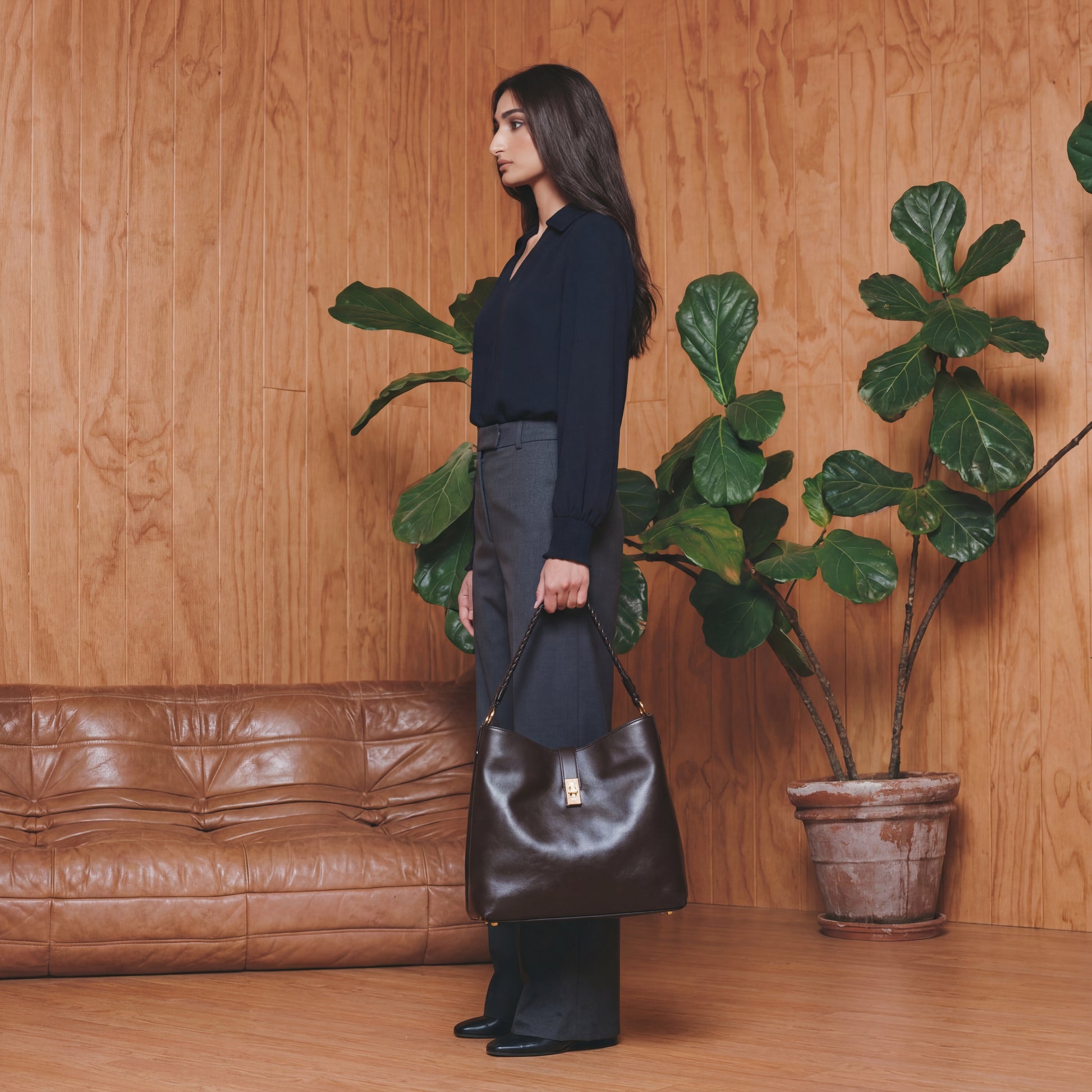 Woman holding a brown leather handbag in a room with wooden walls and a potted plant.