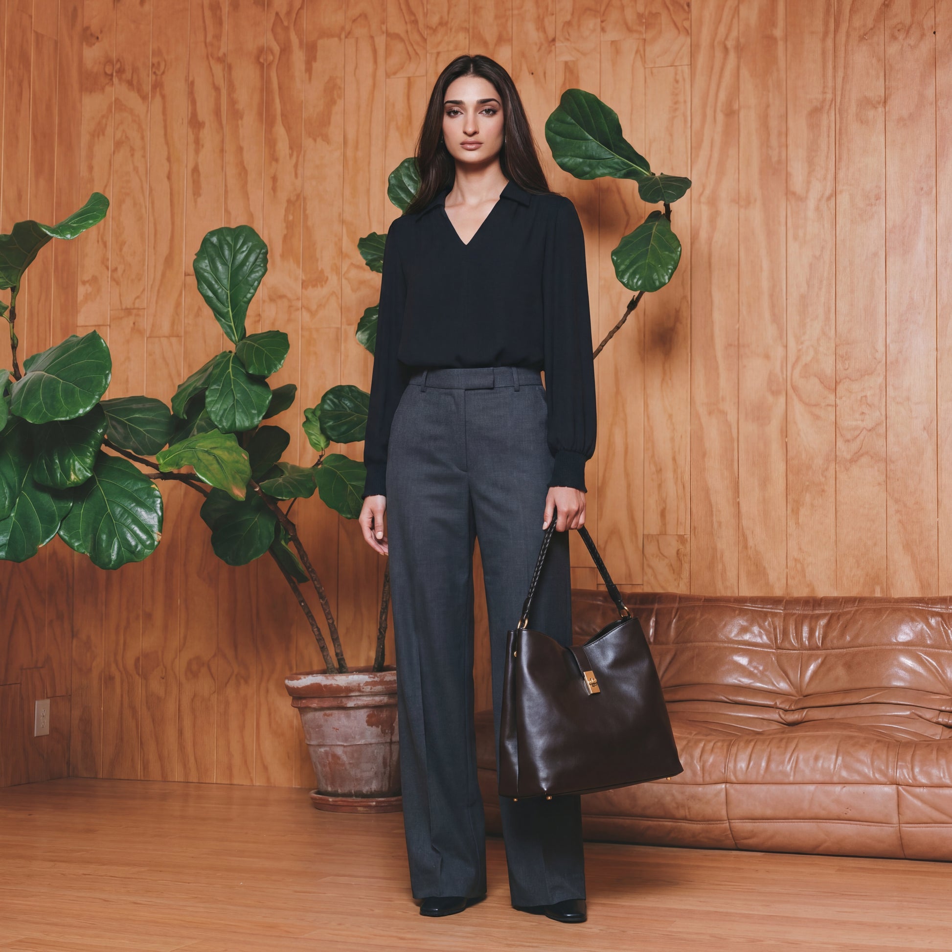 Woman in black top and grey pants holding a black handbag in front of a wooden wall with plants and a brown leather couch.