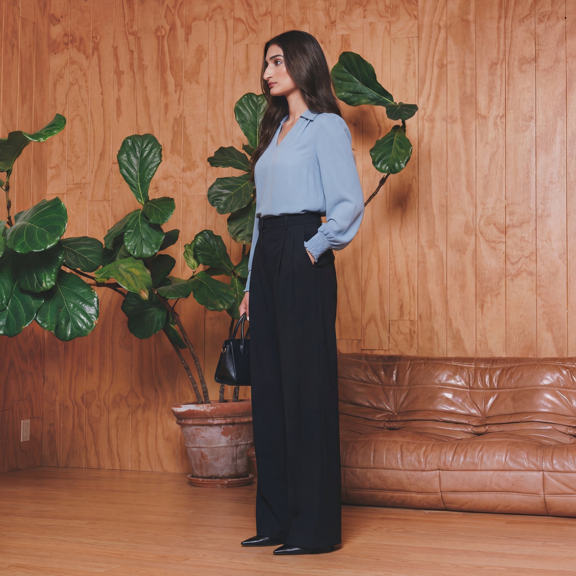 Woman in a light blue shirt and black pants standing in a room with wooden walls, a brown leather couch, and large green plants.