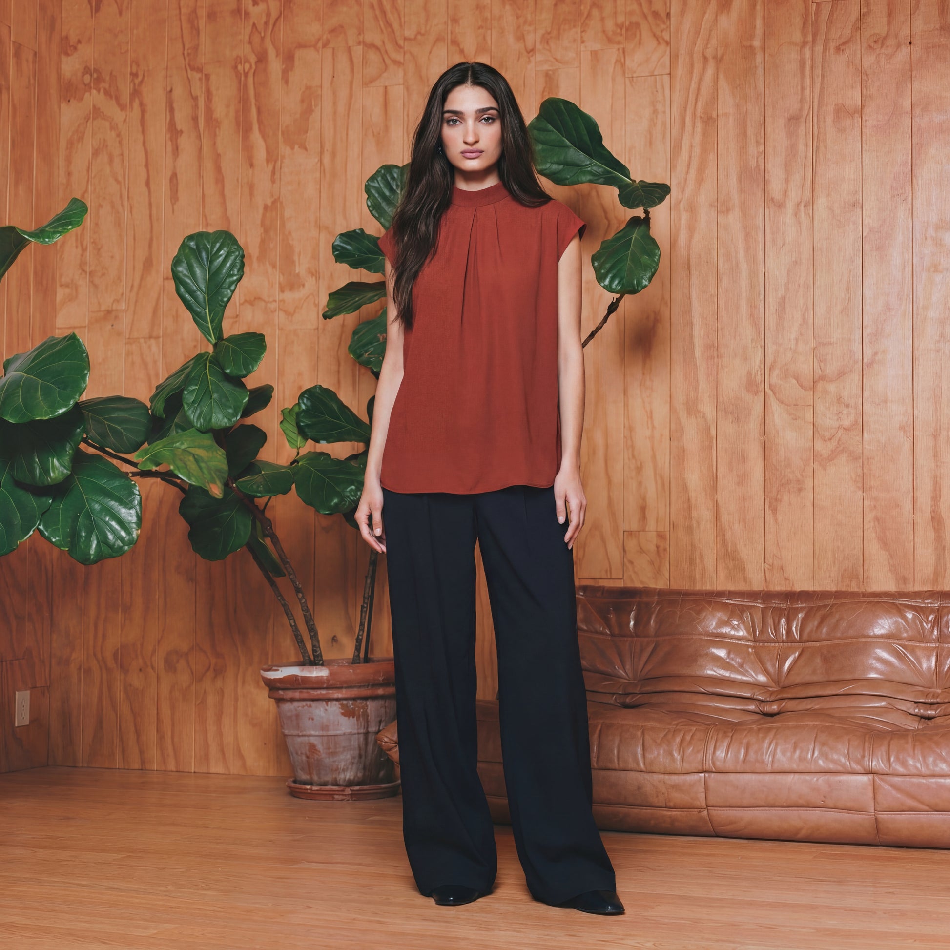 Woman in a rust-colored top and black pants standing in a room with wooden walls, a brown leather couch, and a potted plant.