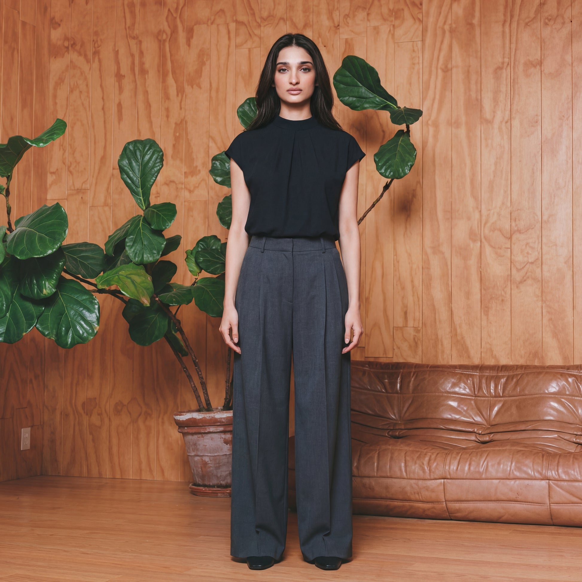 Woman in black top and grey pants standing in a room with wooden walls, a brown leather couch, and potted plants.