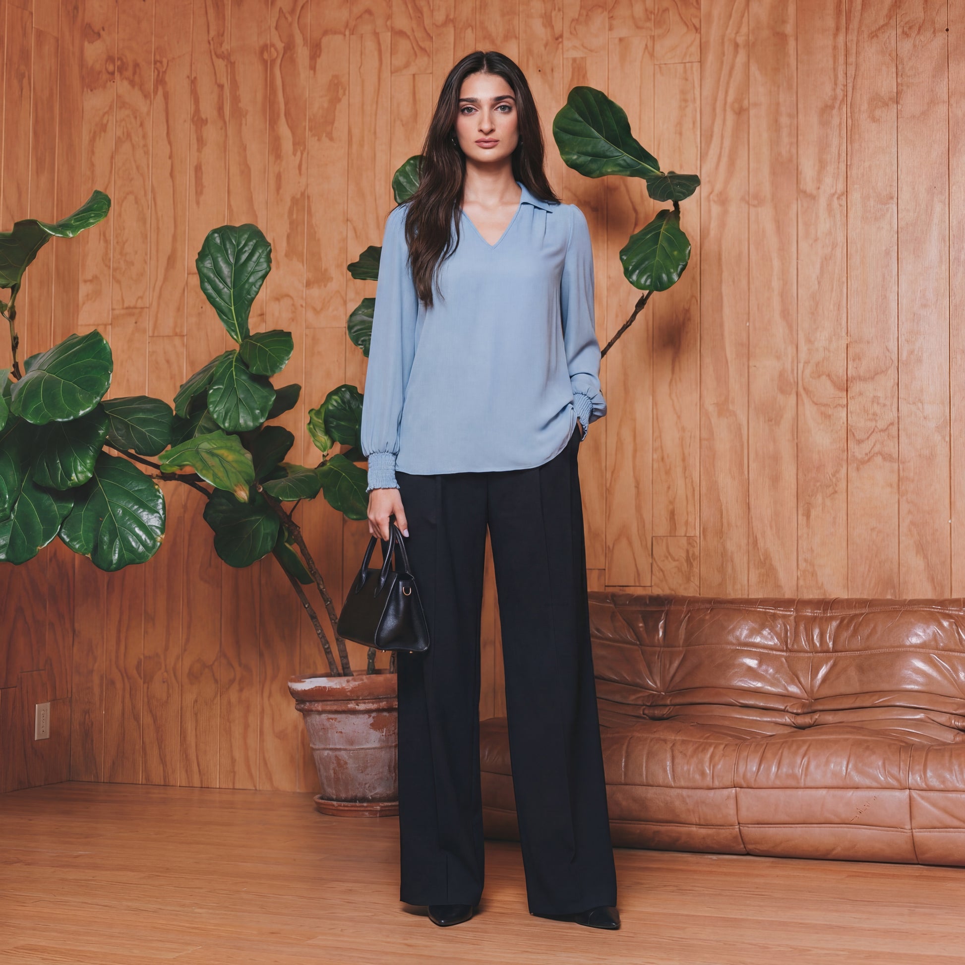 Woman in a blue blouse and black pants standing in front of a wooden wall with plants and a brown leather couch.