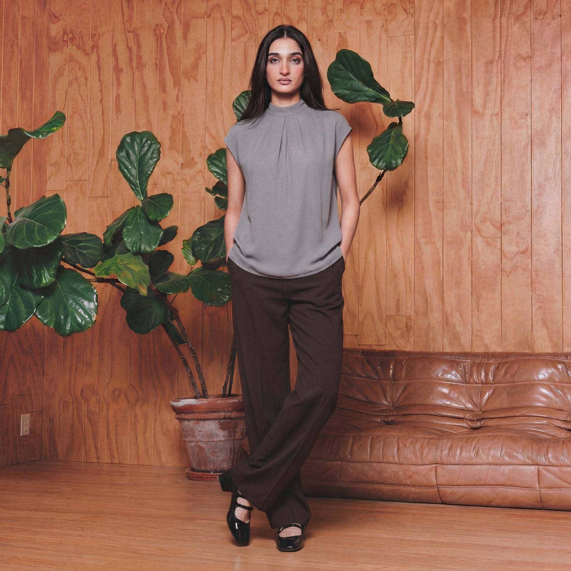 Woman standing in a room with wooden walls, a brown leather couch, and potted plants.