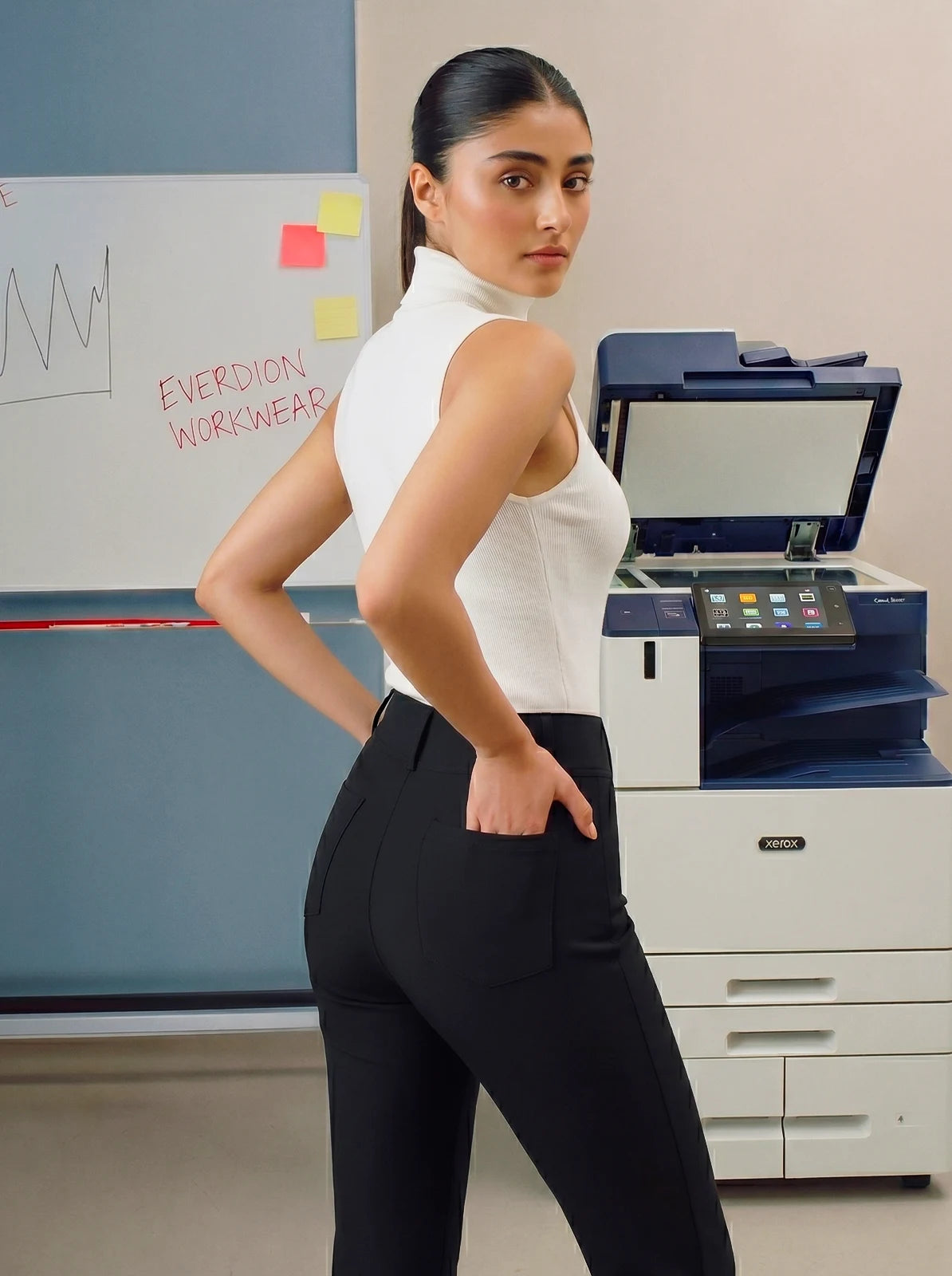 Woman in a white top and black pants standing next to a printer in an office setting.