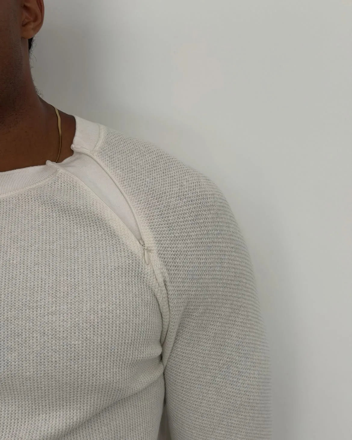 Close-up of a person wearing a white textured knit long-sleeve top against a plain light grey wall.