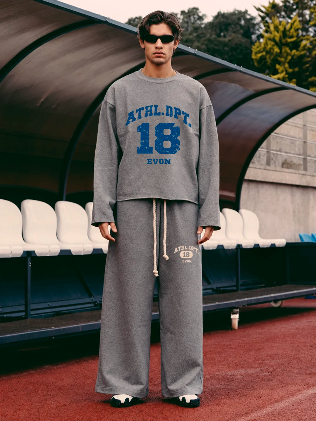 A man wearing a gray athletic tshirt and matching sweatpants stands on a red track field under a dugout structure. The sweatshirt has blue collegiate-style text reading 'ATHL.