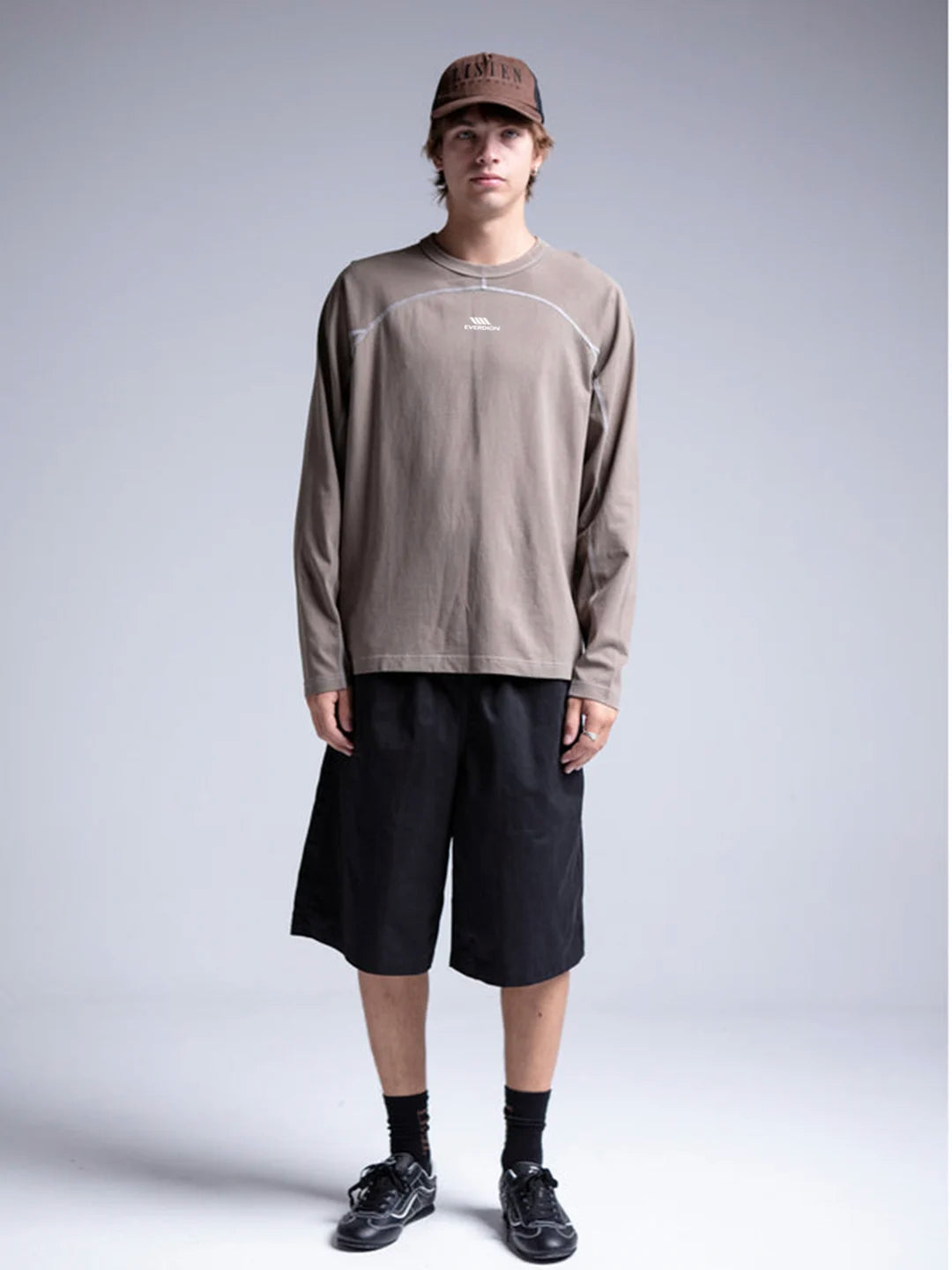 A person wearing a taupe-brown long-sleeve crewneck shirt with white piping details, black shorts, black socks, and black sneakers, along with a brown baseball cap.