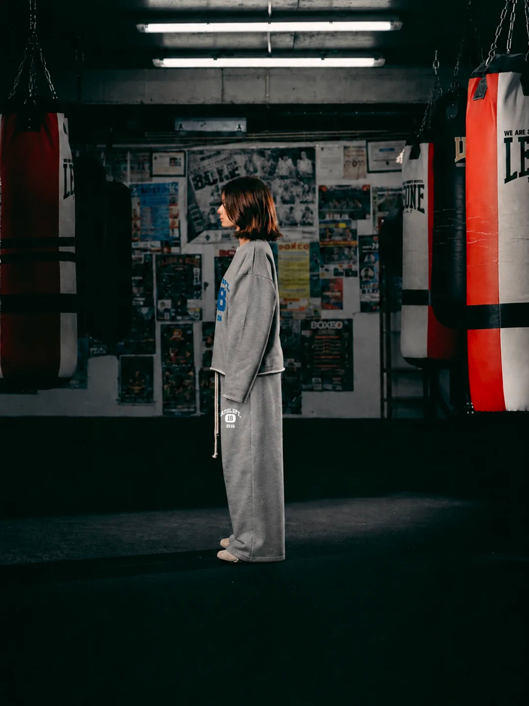 A person wearing a loose grey martial arts uniform or robe with white trim and a white belt. Heavy punching bags hang on both sides, and a wall covered in posters is behind them.