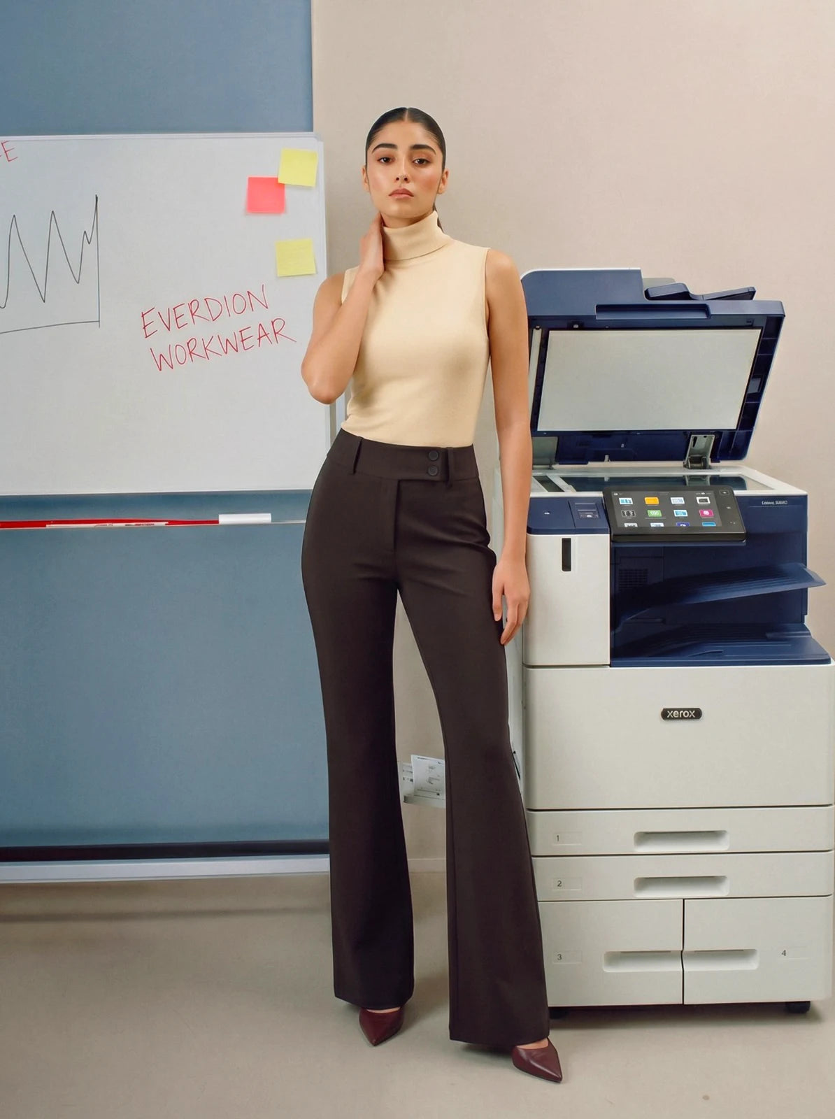 Person wearing brown pants standing in an office setting with a printer and whiteboard in the background.