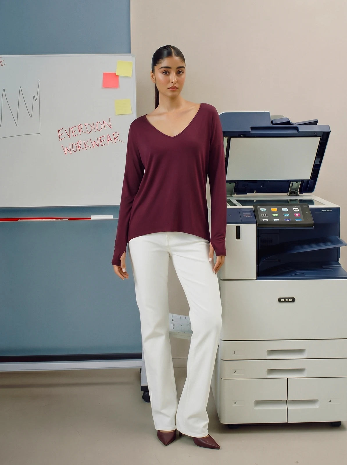 Woman wearing a maroon long-sleeve shirt and white pants standing in an office setting.