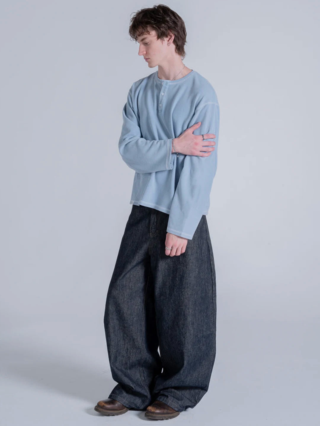 A model wearing a light blue long-sleeve henley t-shirt and very wide-leg dark grey or black denim pants