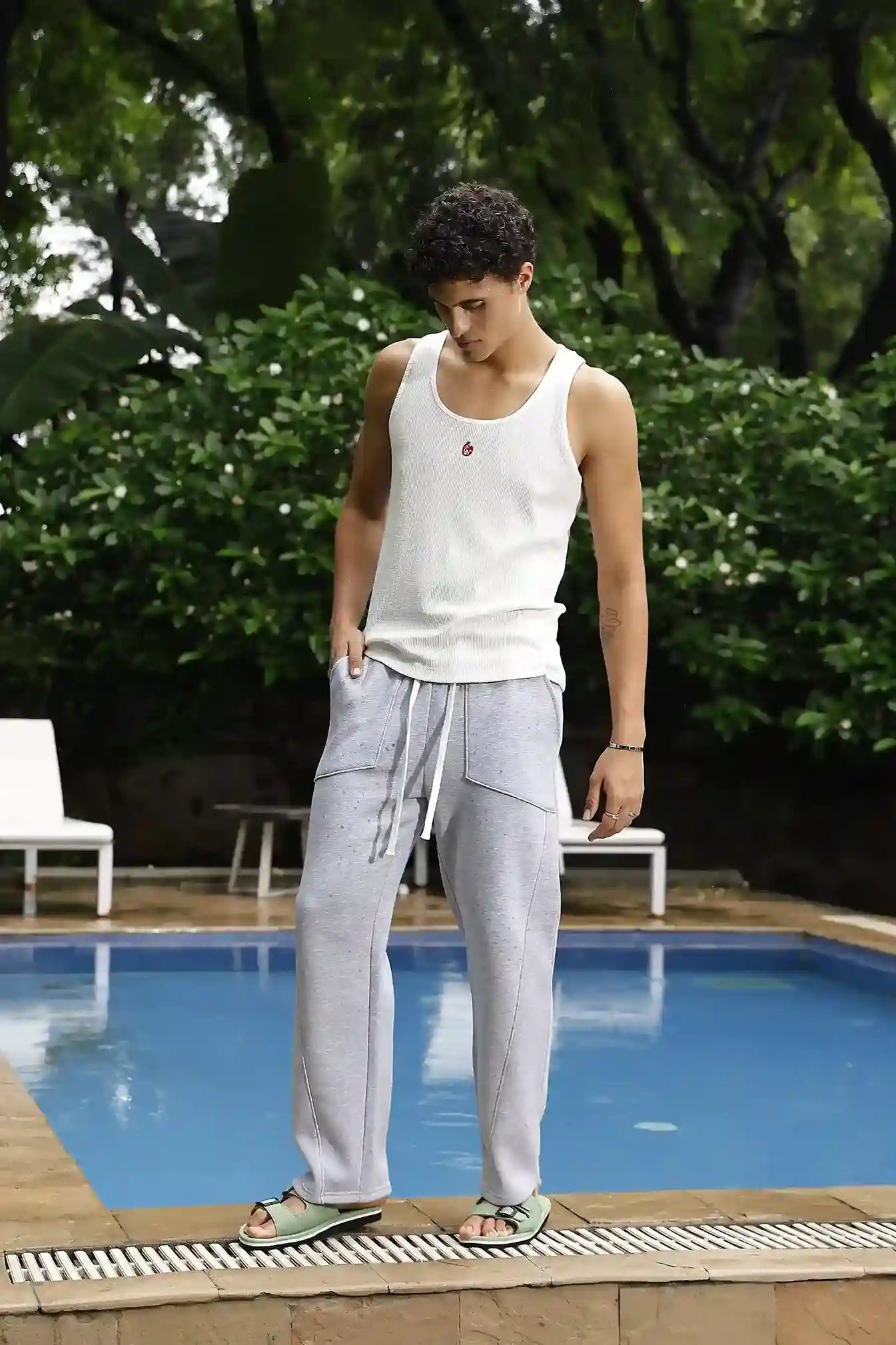 Man in a white tank top and gray pants standing by a pool with greenery in the background