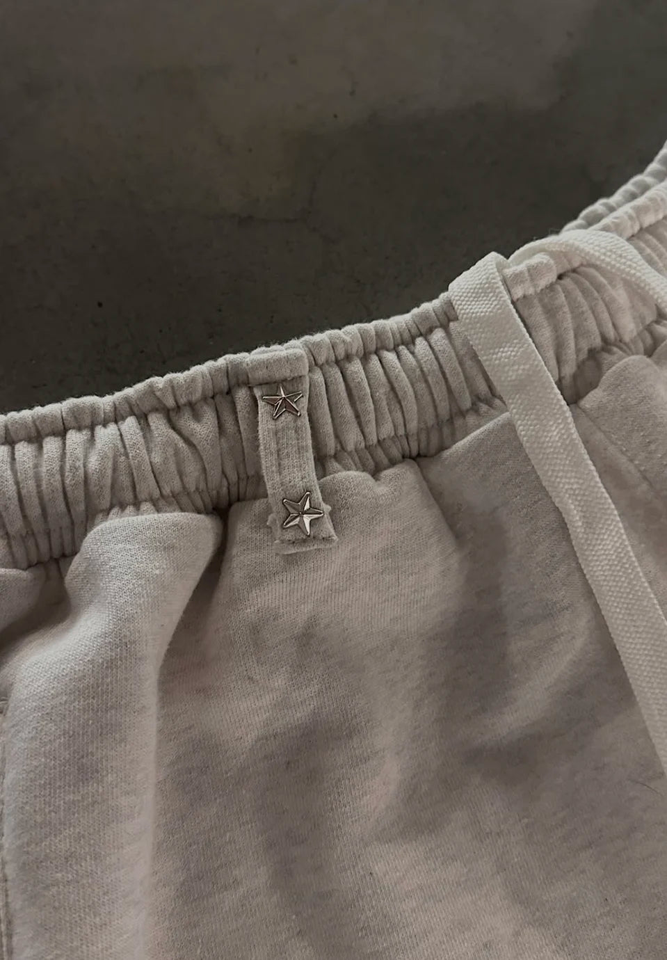 Close-up of a light beige or off-white fabric garment, showing a gathered elastic waistband with a small embroidered star detail and a drawstring.