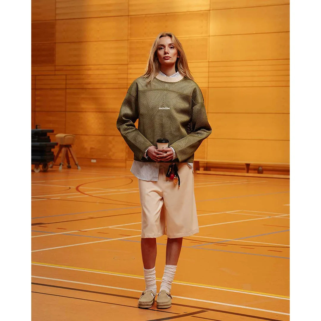 Person wearing a green sweater and beige shorts standing on a wooden floor.