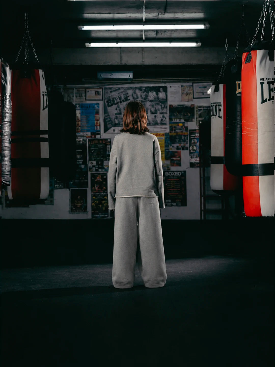 A person wearing a light grey sweatshirt and matching wide-leg sweatpants. Heavy punching bags and a poster-covered wall are visible in the background.