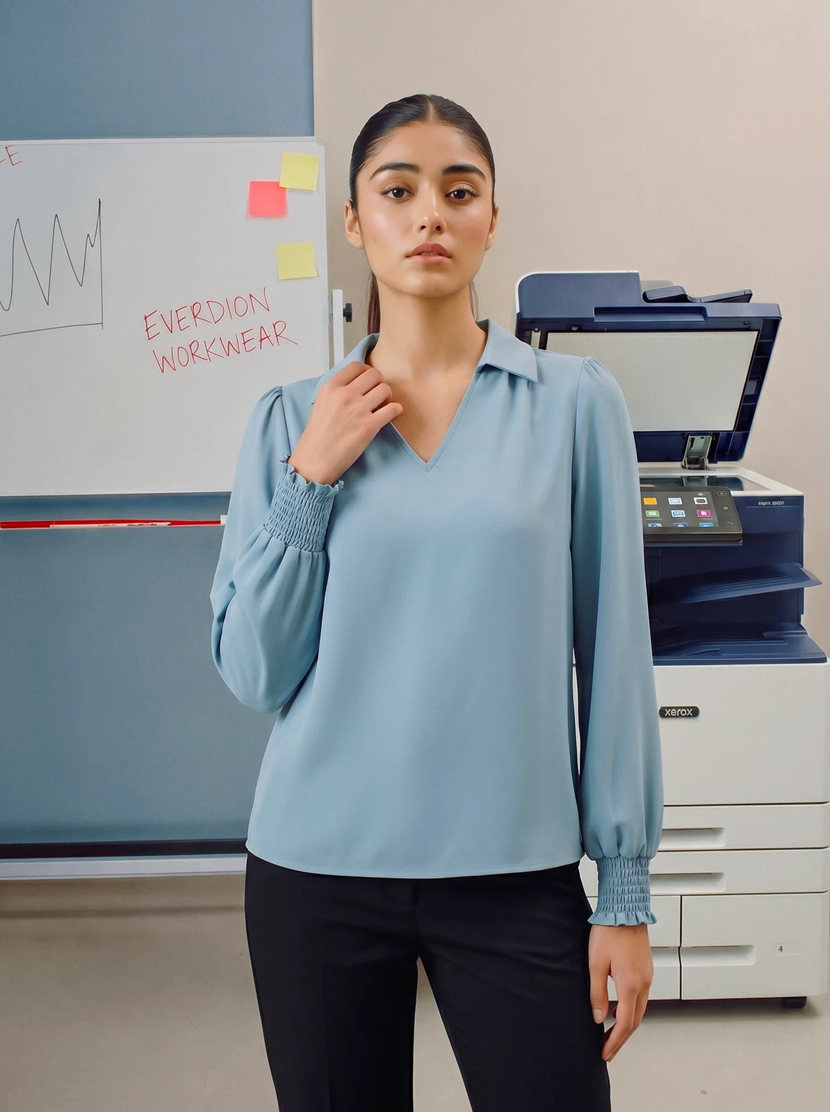 Person wearing a light blue blouse standing in an office setting with a printer and whiteboard in the background.