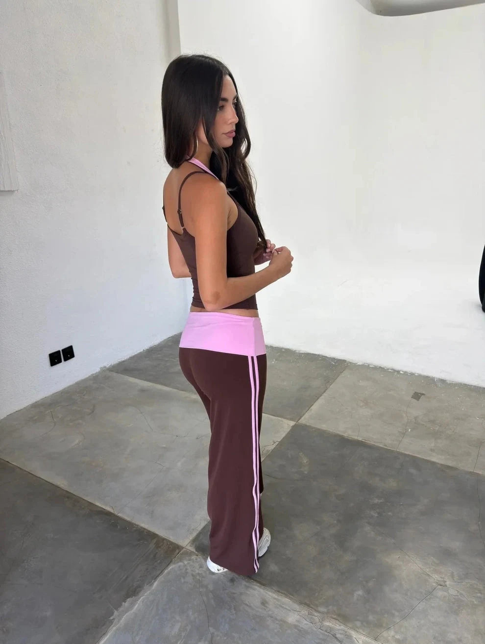 A woman wearing a matching athletic set consisting of a dark brown halter crop top and wide-leg pants with a bright pink waistband and white side stripes, standing in a minimalist white-walled room with concrete floors.