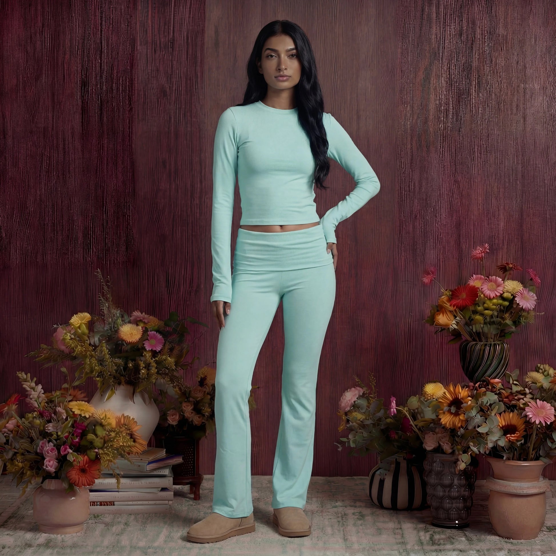 Woman in a light blue cotton fold over pants and top standing among potted flowers against a wooden wall.