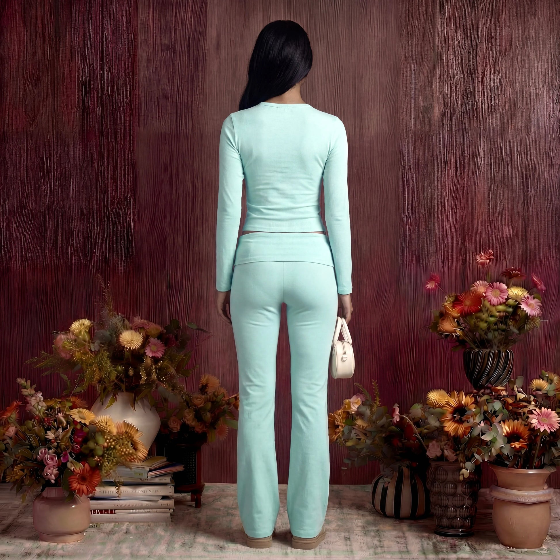 Woman in a light blue cotton fold over pants and top standing among potted plants against a wooden wall.