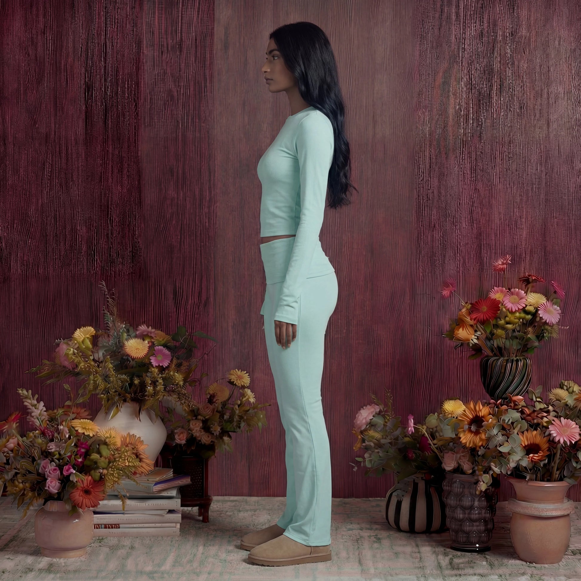 Woman in a light green cotton fold over pants and top standing among potted flowers against a textured purple wall.