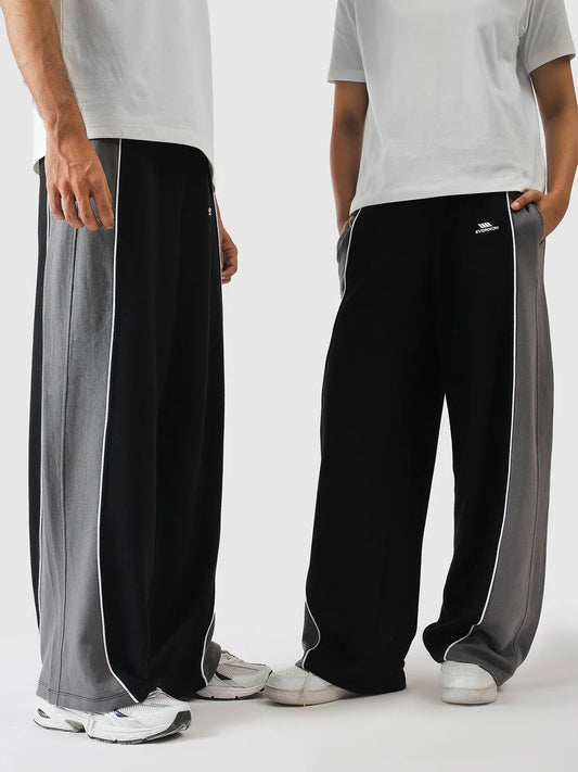 Two models wearing wide-leg black sweatpants with grey side panels and white piping, paired with white t-shirts and sneakers