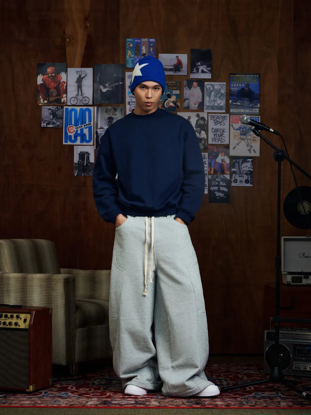 Person wearing a blue sweatshirt and light grey sweatpants standing in a room with posters on the wall.