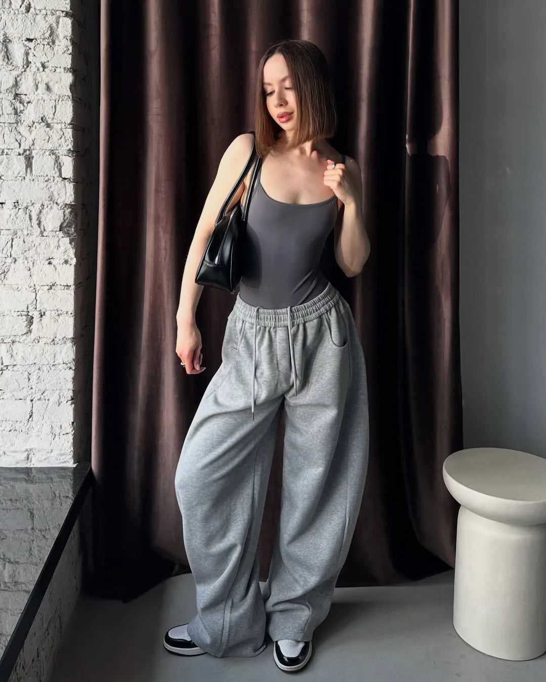 Woman in grey sweatpants standing against a brown curtain.