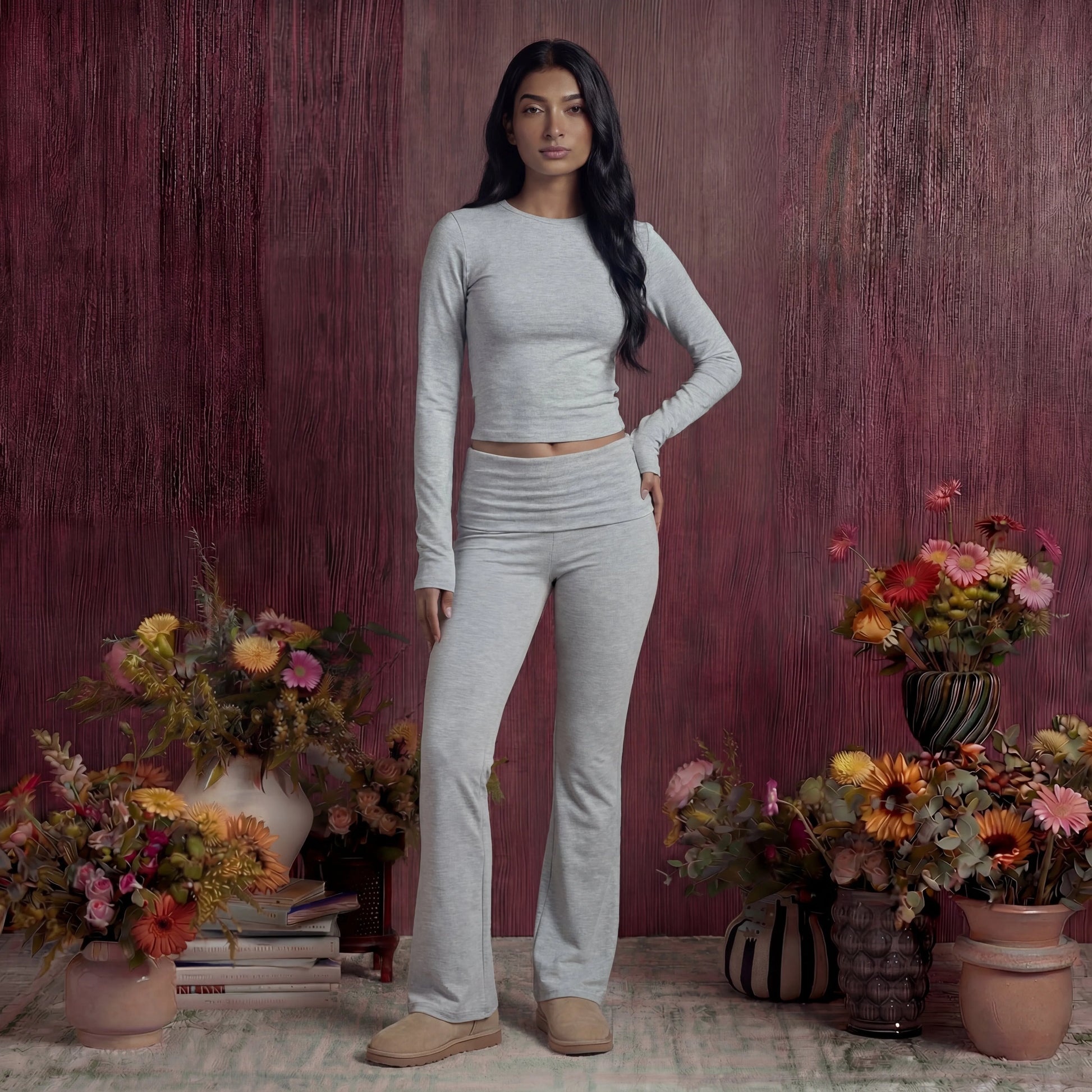 Woman in grey cotton fold over pants and top standing among potted flowers against a textured purple wall.